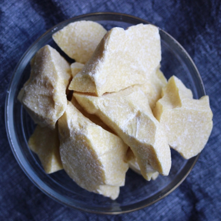Chunks of solid cocoa butter in a clear glass bowl on a blue textured surface.